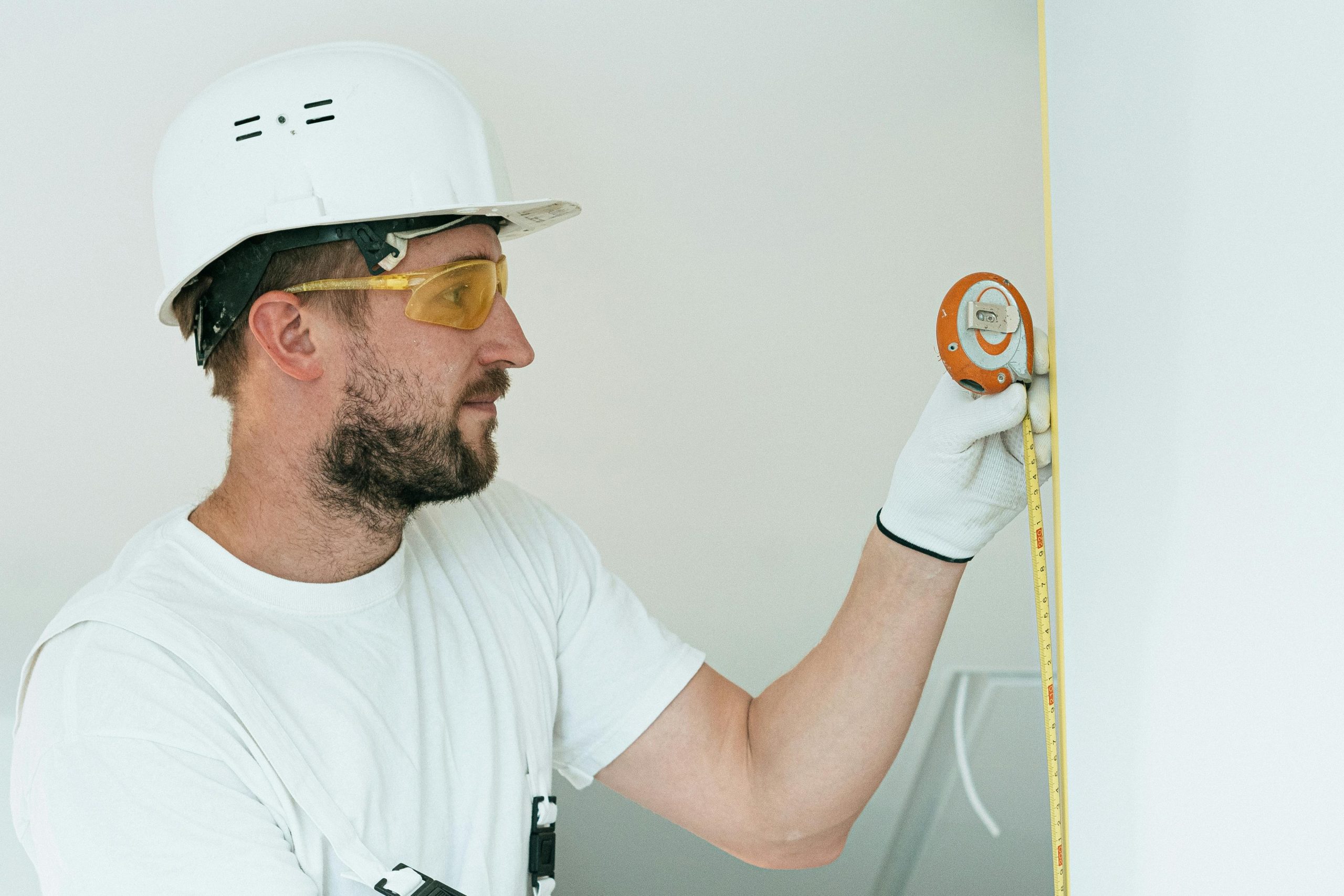 man wearing a white hard hat using a tape measure to check the length of a wall